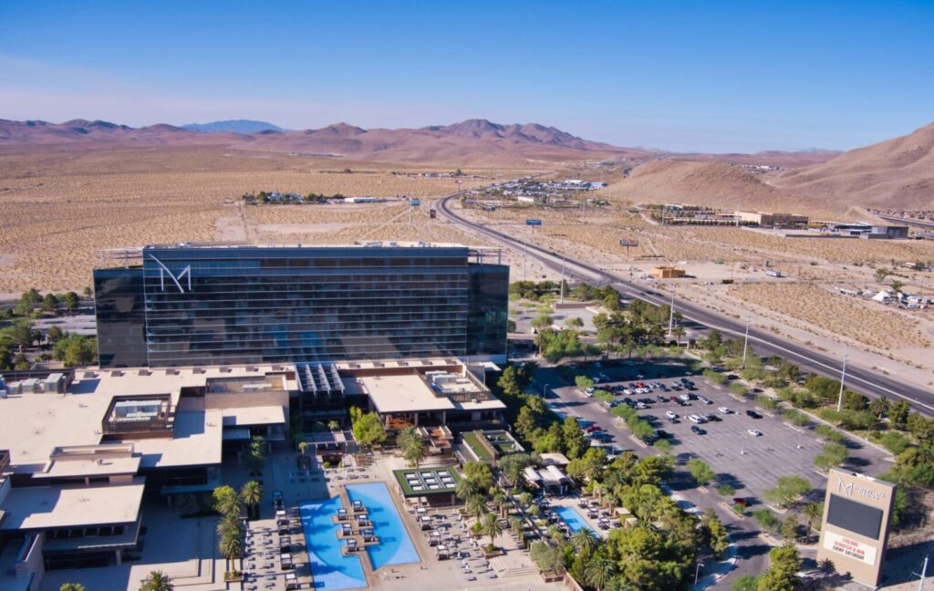 Aerial view of a large modern hotel and casino complex surrounded by desert landscape, with mountains in the distance, a parking lot, swimming pools, and a highway running nearby under a clear blue sky.