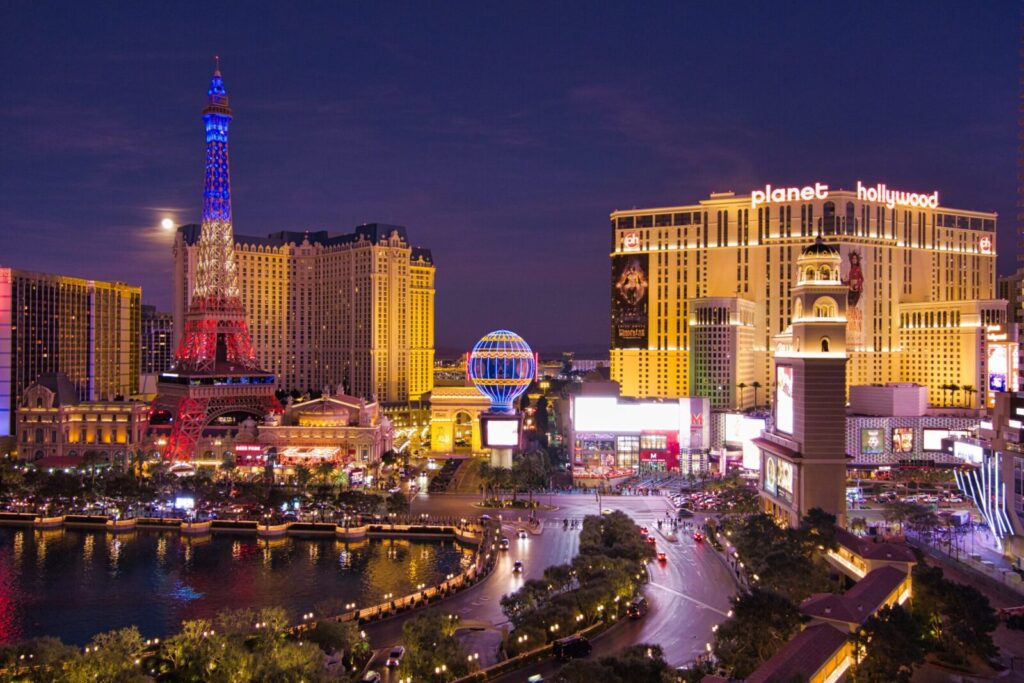 A nighttime view of the Las Vegas Strip captured through drone photography in Las Vegas showcases the illuminated Eiffel Tower replica, vibrant hotels, neon signs, bustling streets with cars and palm trees beneath a glowing deep blue sky.