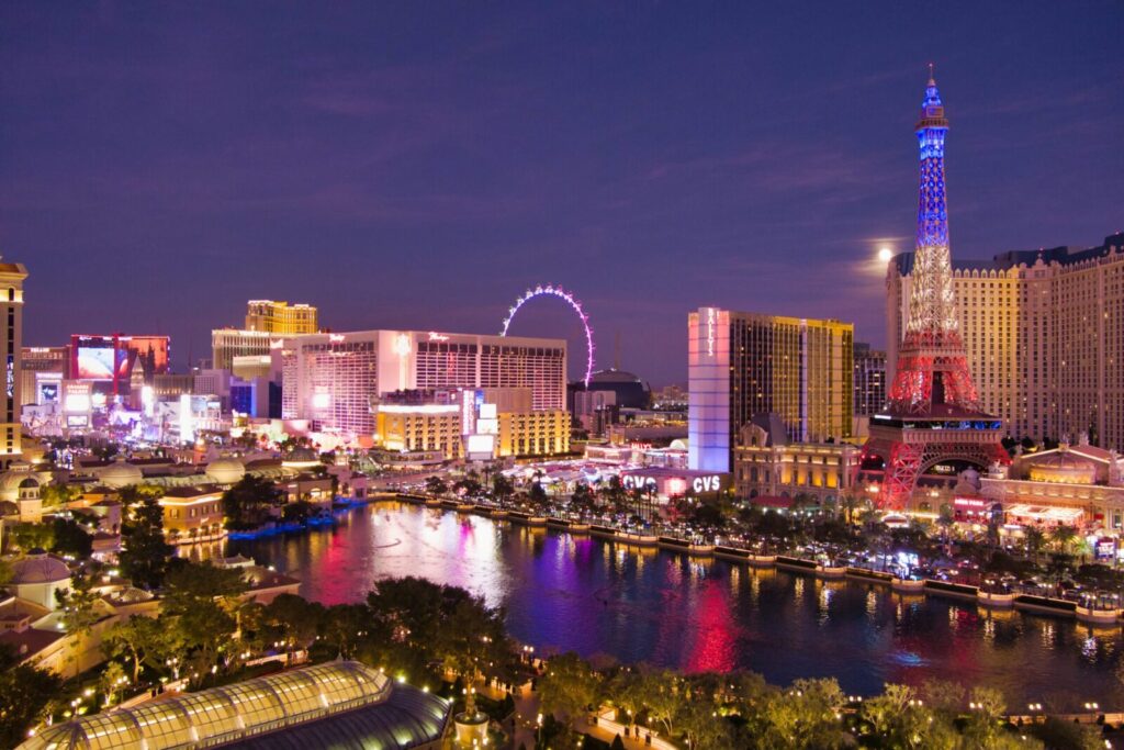 A nighttime view of the Las Vegas Strip, captured by drone photography in Las Vegas, features bright lights, colorful hotels, a replica of the Eiffel Tower, and a large Ferris wheel reflected in the water below.