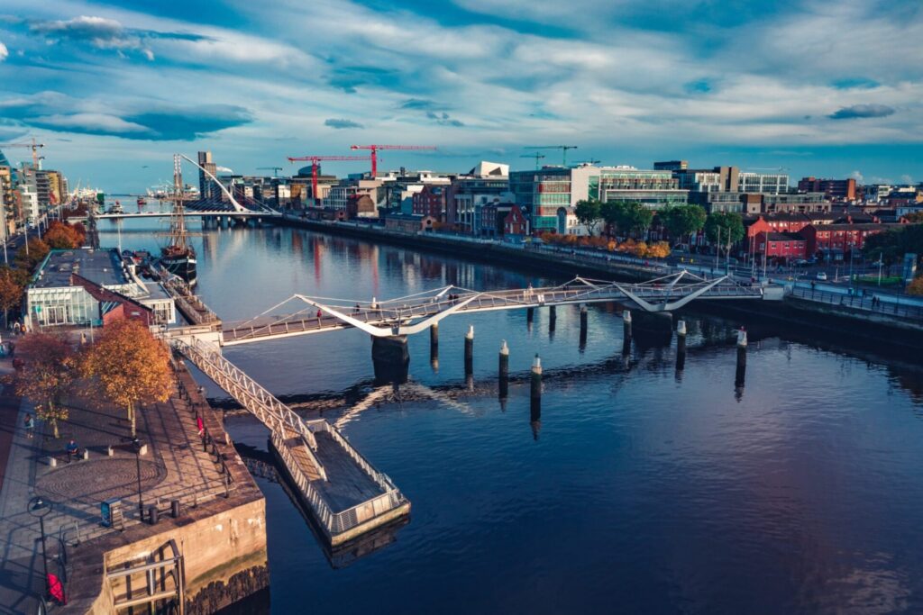 Aerial view captured through drone photography: a modern white pedestrian bridge spans a calm river, surrounded by city buildings, autumn trees, cranes, and a partly cloudy blue sky.
