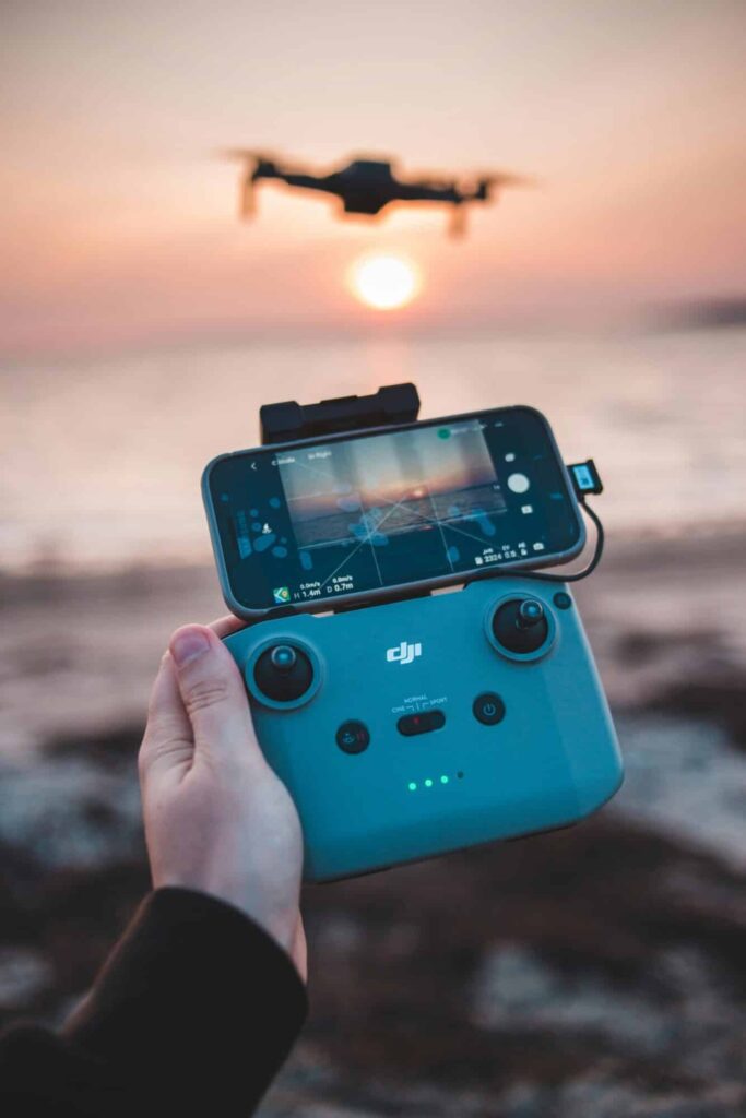 A hand holds a DJI drone remote controller with a smartphone attached, displaying a live feed of a Drone Photography Company-Sky View Las Vegas drone flying over a beach at sunset. The blurred drone and sun glow in the background.