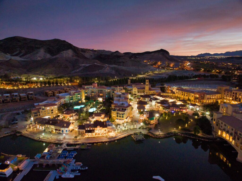 Aerial view of a brightly lit Lake Las Vegas lakeside resort town at dusk, surrounded by mountains, with boats docked at a marina and glowing buildings reflecting in the calm water—Elevate Your Views with a Drone Photography Company.