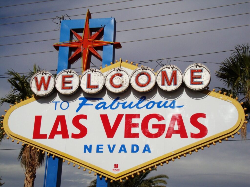 The famous Welcome to Fabulous Las Vegas, Nevada sign with bold red, blue, and yellow letters, outlined by lights, set against palm trees and a partly cloudy sky—a classic scene often captured in Las Vegas aerial photography.