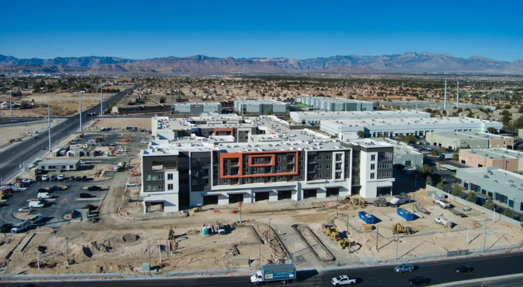 Aerial view of a modern building under construction, with dirt lots, vehicles, and equipment on site. Industrial buildings and mountains appear in the distance under a clear sky—showing why How to Choose the Right Drone Service for Construction Projects matters.