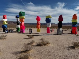 Seven Magic Mountains-Colorful stacked boulders form several tall columns in a desert landscape near Las Vegas. People walk around, take photos, and occasionally spot Las Vegas drones flying under a blue sky with wispy clouds and distant mountains.