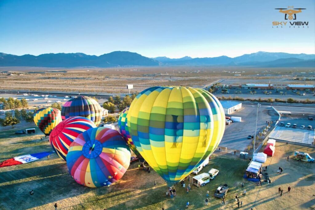 A group of colorful hot air balloons prepare for takeoff on an open field near a road, with mountains and clear blue sky in the background. People and vehicles gather as if attending a vibrant outdoor gallery.