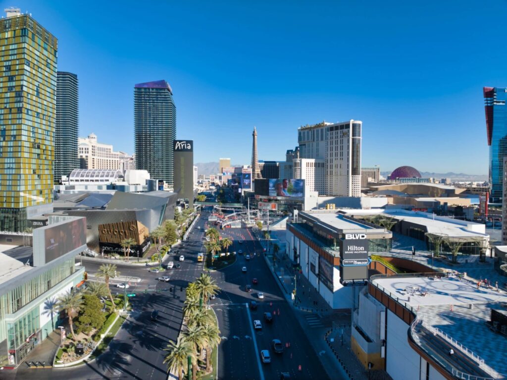Aerial view of the Las Vegas Strip with tall hotels, casinos, palm trees, and busy streets under a bright blue sky. Catch Vegas From Above: Drone Pics! showcasing the Eiffel Tower replica and dazzling billboards in the distance.