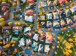 Aerial view of a suburban neighborhood with houses, streets, green lawns, and trees. Showcasing trends in aerial photography, the image captures a winding road and homes amid autumn foliage in various shapes and sizes.