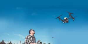 A man wearing sunglasses and a plaid shirt operates a flying drone with a remote control under a clear blue sky. Trees and a light pole are visible in the background.