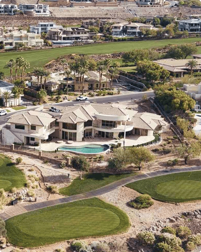 Aerial view of a large, modern house with a pool, surrounded by landscaped greenery and located near a golf course, with other luxury homes and rolling hills in the background.