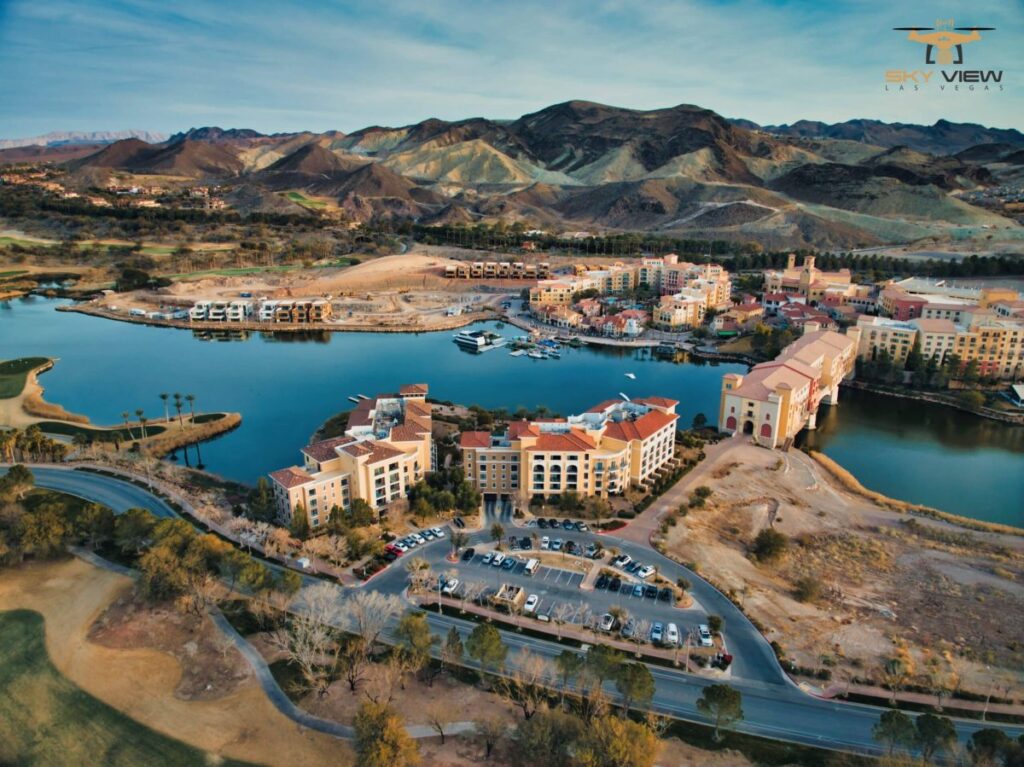 Aerial view of Lake Las Vegas resort with Mediterranean-style buildings, surrounded by mountains, golf courses, winding roads, and sparse trees under a partly cloudy sky—captured with professional drone photography services.