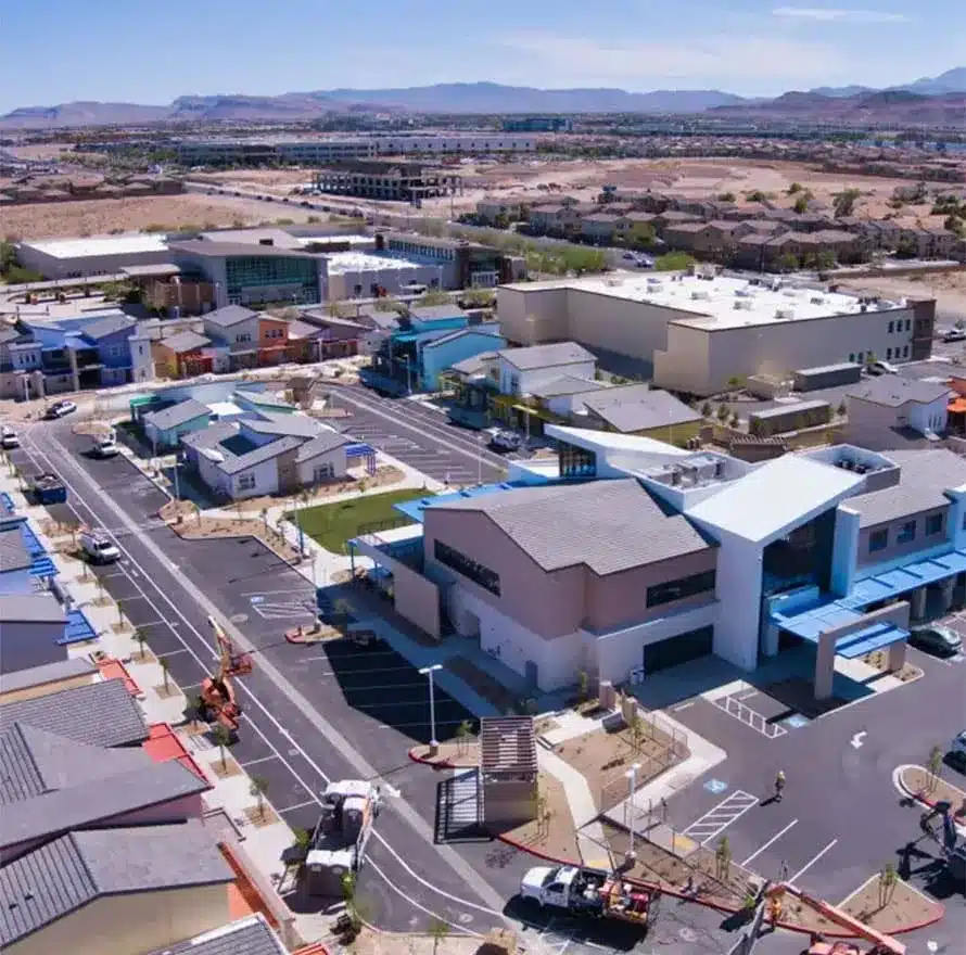 Aerial view of a modern suburban complex with multiple buildings, parking lots, and landscaped areas, surrounded by roads and distant mountains under a clear sky.