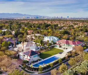 Aerial view of a luxury suburban neighborhood with large houses, manicured lawns, a swimming pool, and tree-lined streets; city skyline and mountains visible in the background under a clear sky.
