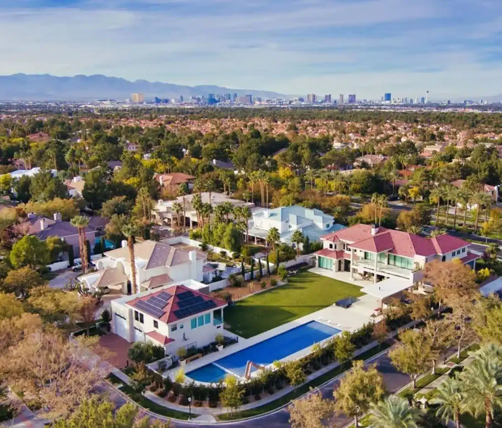 Aerial view of a luxury suburban neighborhood with large houses, manicured lawns, a swimming pool, and tree-lined streets; city skyline and mountains visible in the background under a clear sky.