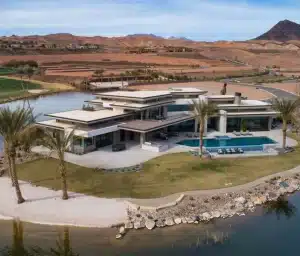 A large, modern mansion with a pool sits on a small island surrounded by water, desert landscaping, palm trees, and distant mountains under a clear sky.