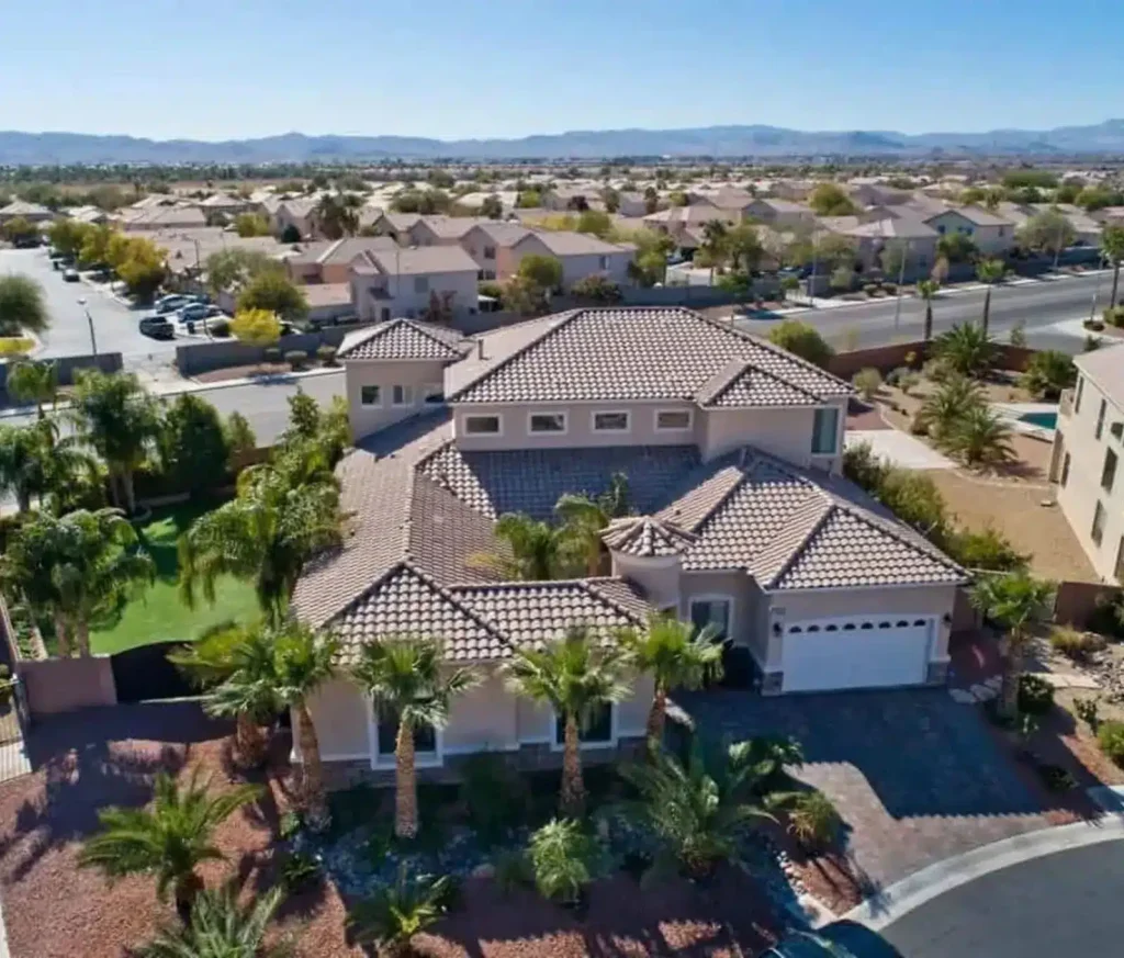 Aerial view of a large, modern house with a tile roof, palm trees, and landscaped yard in a suburban neighborhood with similar homes and distant mountains under a clear blue sky.