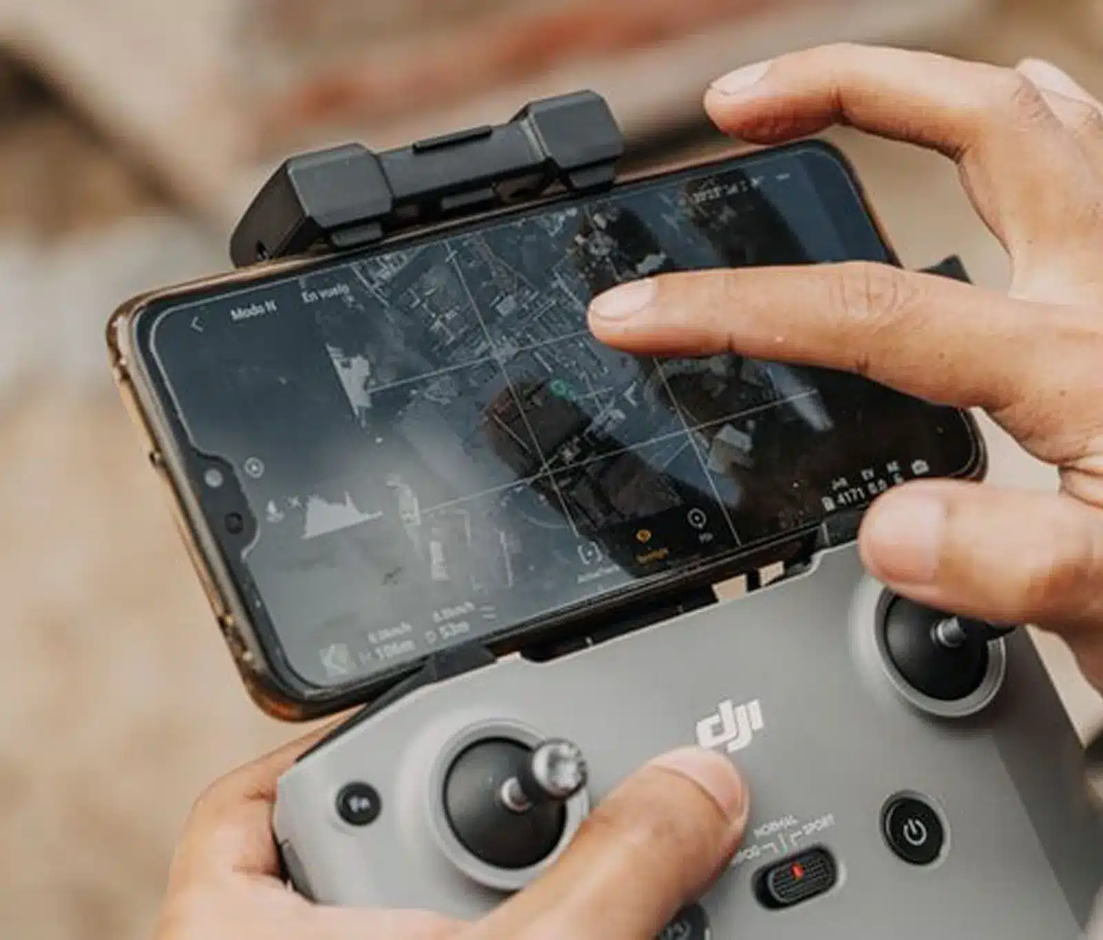 A person operates a DJI drone remote controller while viewing a live aerial map on a smartphone screen attached to the controller.