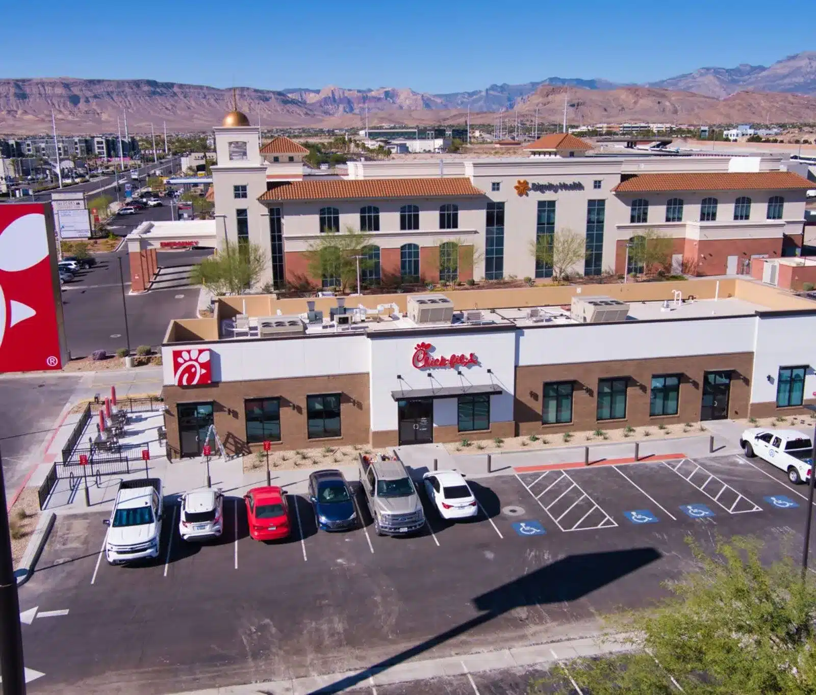 Aerial view of a Chick-fil-A restaurant with a parking lot in front, several cars parked, nearby buildings, and mountains in the background under a clear blue sky.