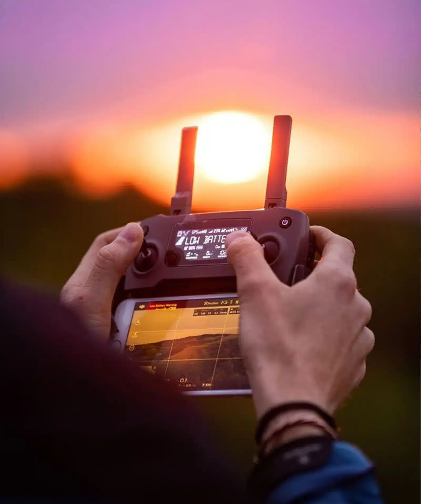 A person holds a remote control with a screen, likely for a drone, at sunset. The glowing sun is framed between the controllers antennas, with warm light and blurred scenery in the background.