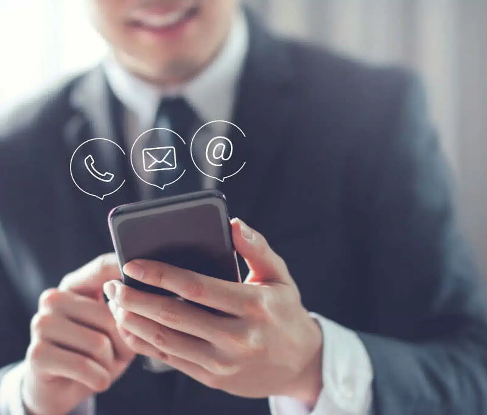 A person in a suit holding a smartphone, with icons for WhatsApp, email, and the @ symbol digitally superimposed, representing digital communication. The background is blurred.