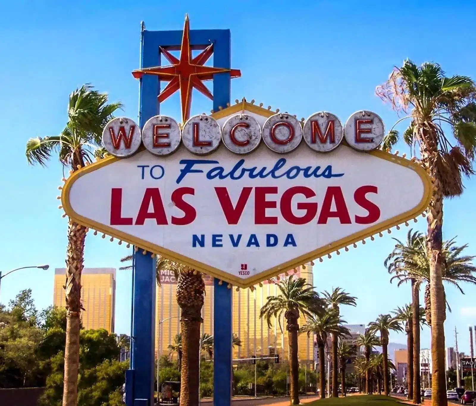 The iconic Welcome to Fabulous Las Vegas Nevada sign stands tall, surrounded by palm trees, with buildings and a clear blue sky in the background.