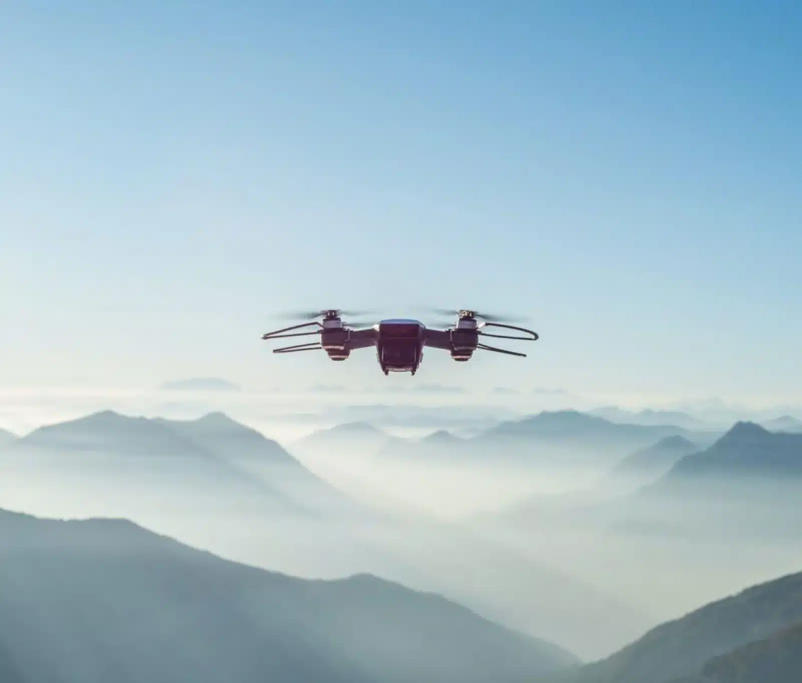 A drone flies above misty mountain ranges under a clear blue sky, capturing a scenic landscape of layered hills and valleys in the early morning light.