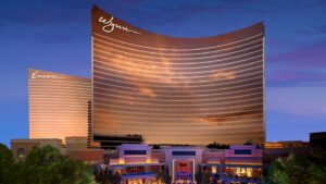 The Wynn and Encore hotel towers in Las Vegas, Nevada, are lit up at dusk with golden reflections on their curved glass facades, standing above a foreground of trees and illuminated shops.